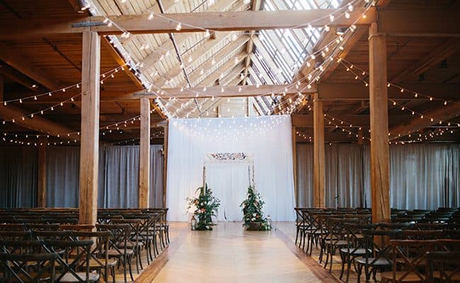 Barn wedding ceremony aisle lit with overhead string lights and a floral arch