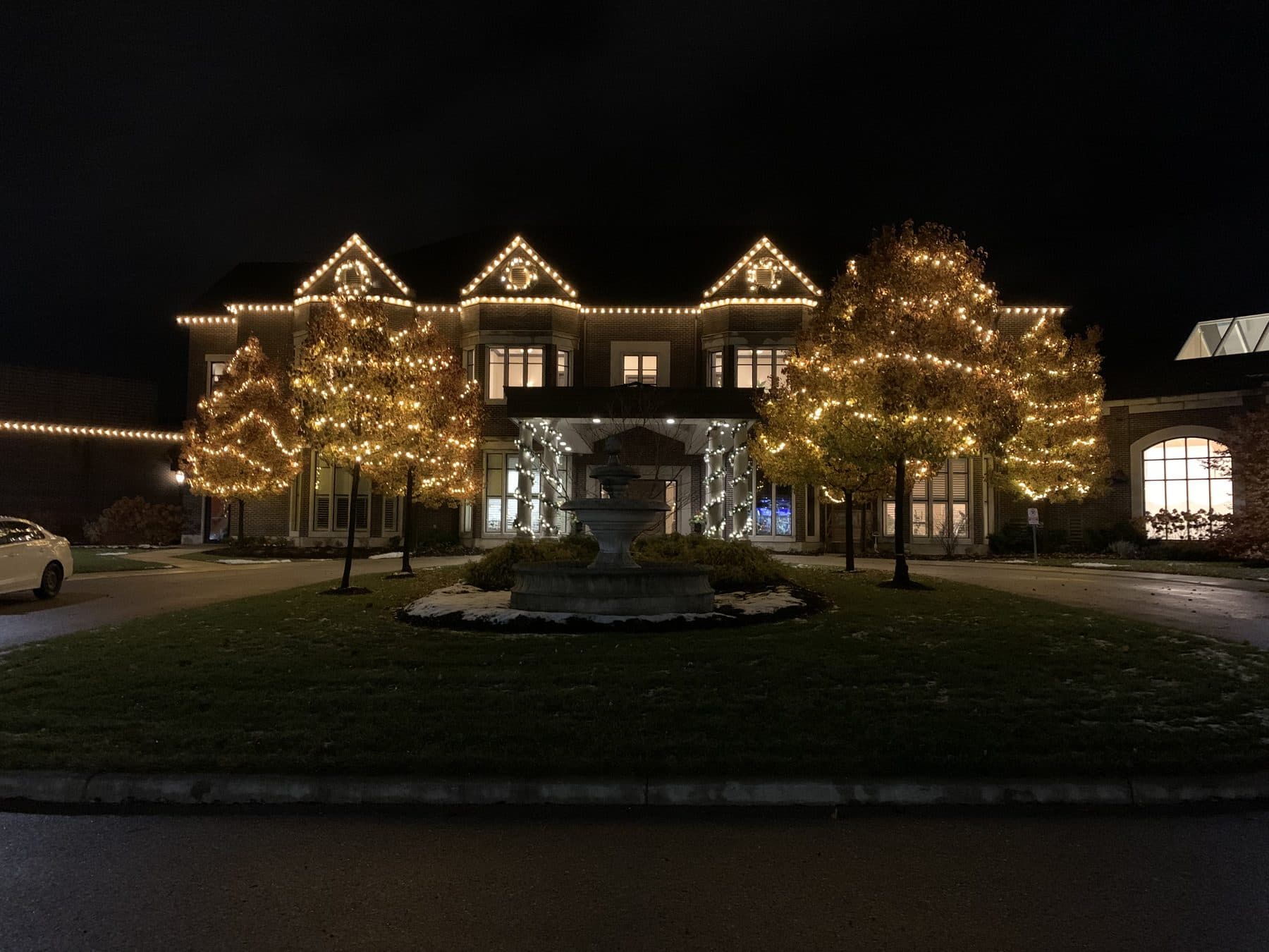 Grand estate venue lit with warm-white roofline lights and wrapped trees around a fountain