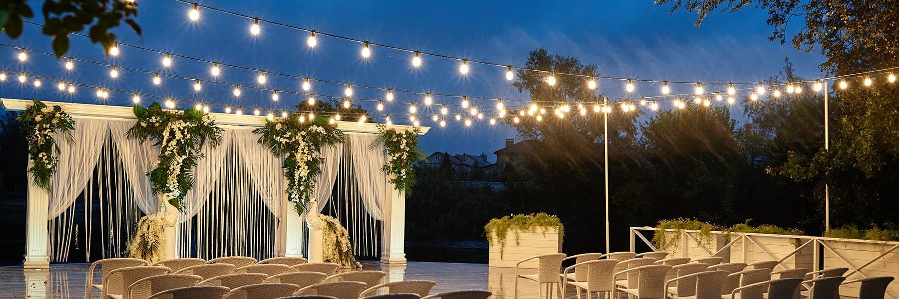 Waterfront wedding ceremony with a draped arch and bistro string lights at blue hour